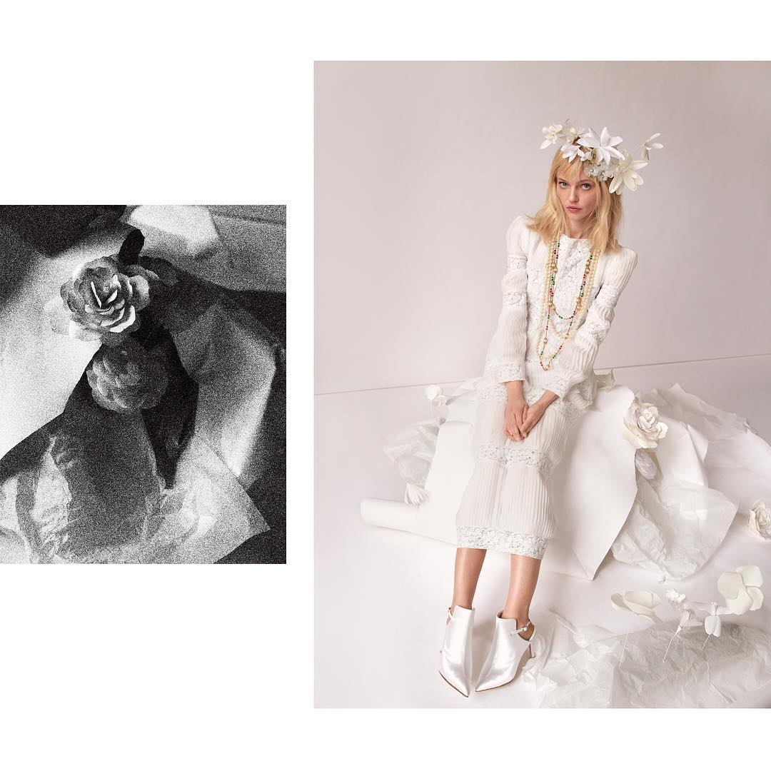 Fashion editorial shot of a blonde woman in white dress and flower crown, seated on white fabric with roses, against a white background.