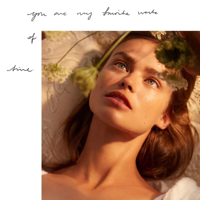 Close-up of a young woman with fair skin and blue eyes gazing upward surrounded by green and white flowers, soft diffused lighting.