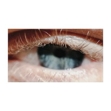 Extreme close-up photograph of a human eye with blue iris, visible eyelashes and surrounding skin, captured in soft, natural light.