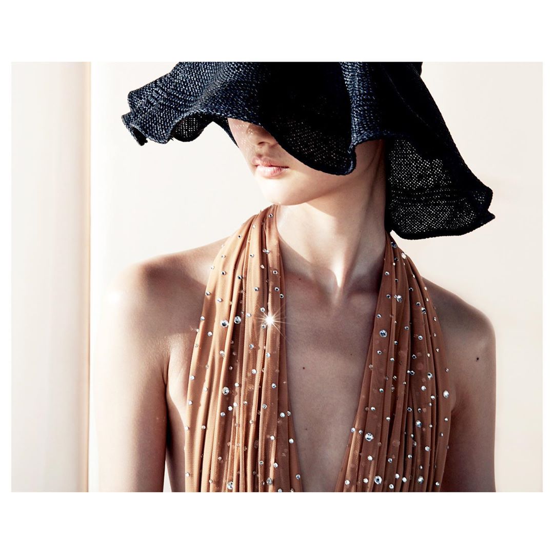 Woman poses in halter top with embellishments and wide-brim hat in an indoor setting with soft, diffused light.