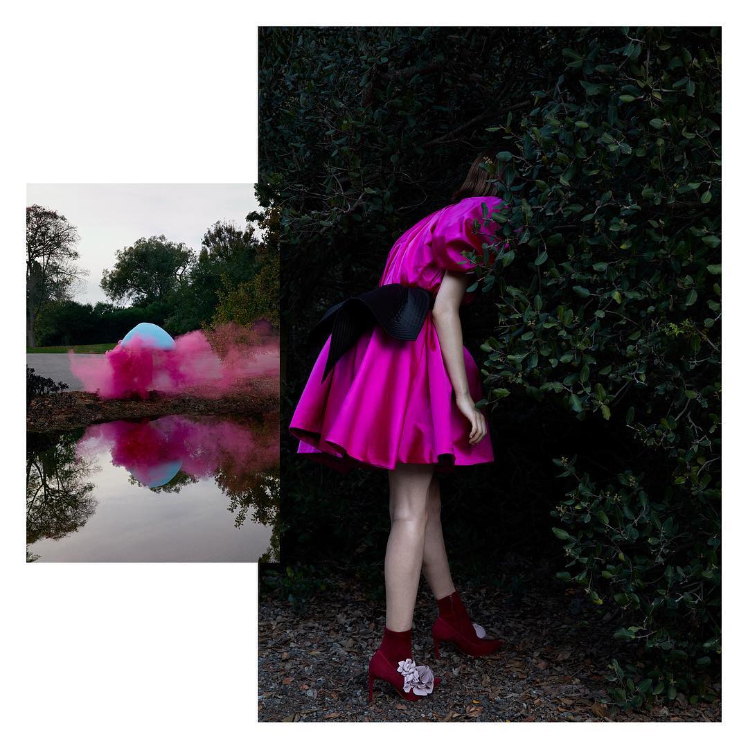 Fashion shot of a woman in a pink dress with a large bow posing in front of green bushes and a blurred image of a dome.