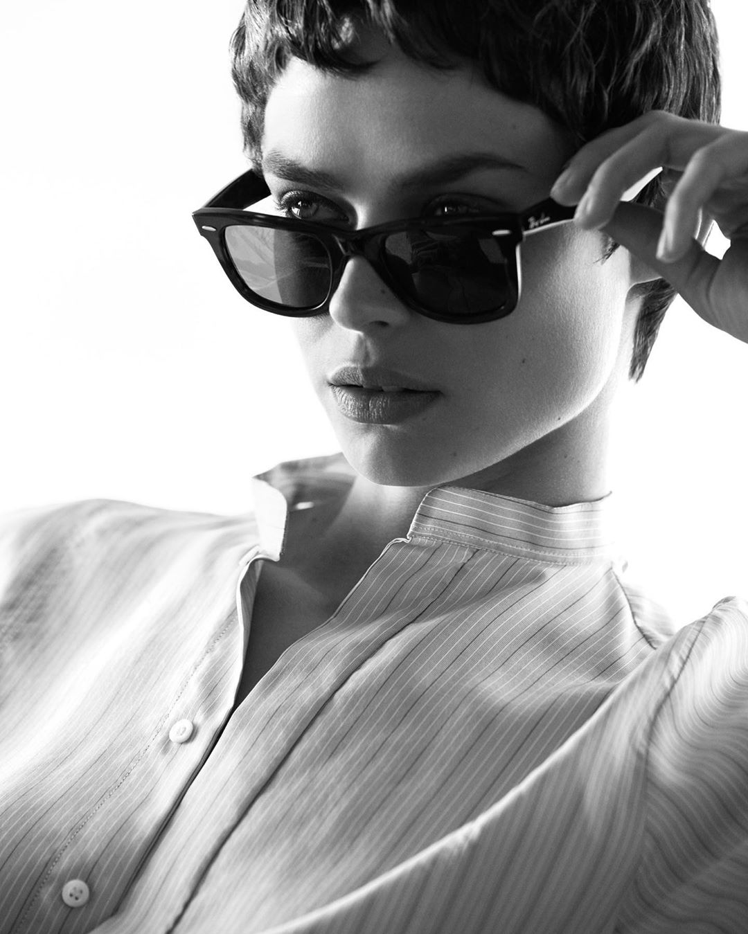Black and white portrait of a young woman with short hair wearing a striped shirt and black Ray-Ban sunglasses, looking away from the camera.