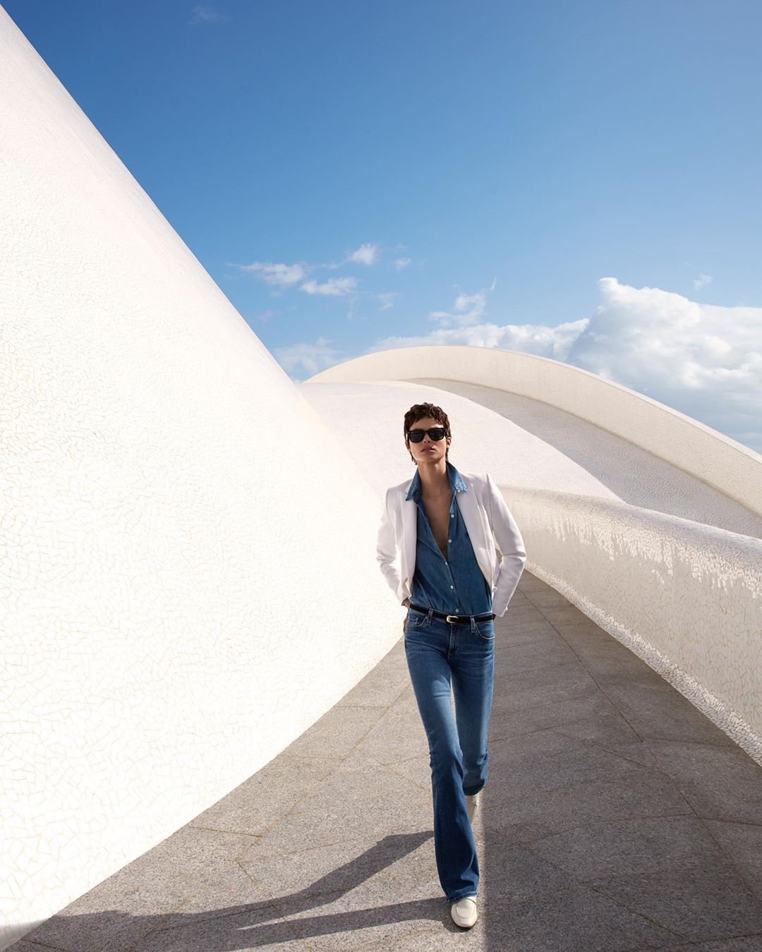 Fashionable woman in flared jeans and white blazer confidently walks along a modern white building exterior under a bright blue sky.