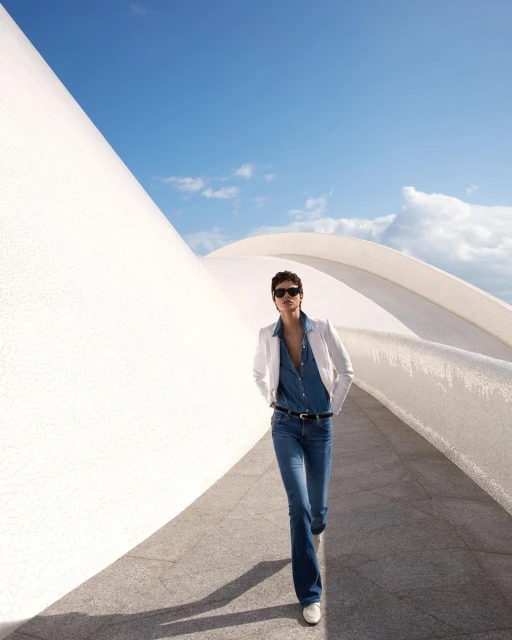 Fashionable woman in flared jeans and white blazer confidently walks along a modern white building exterior under a bright blue sky.