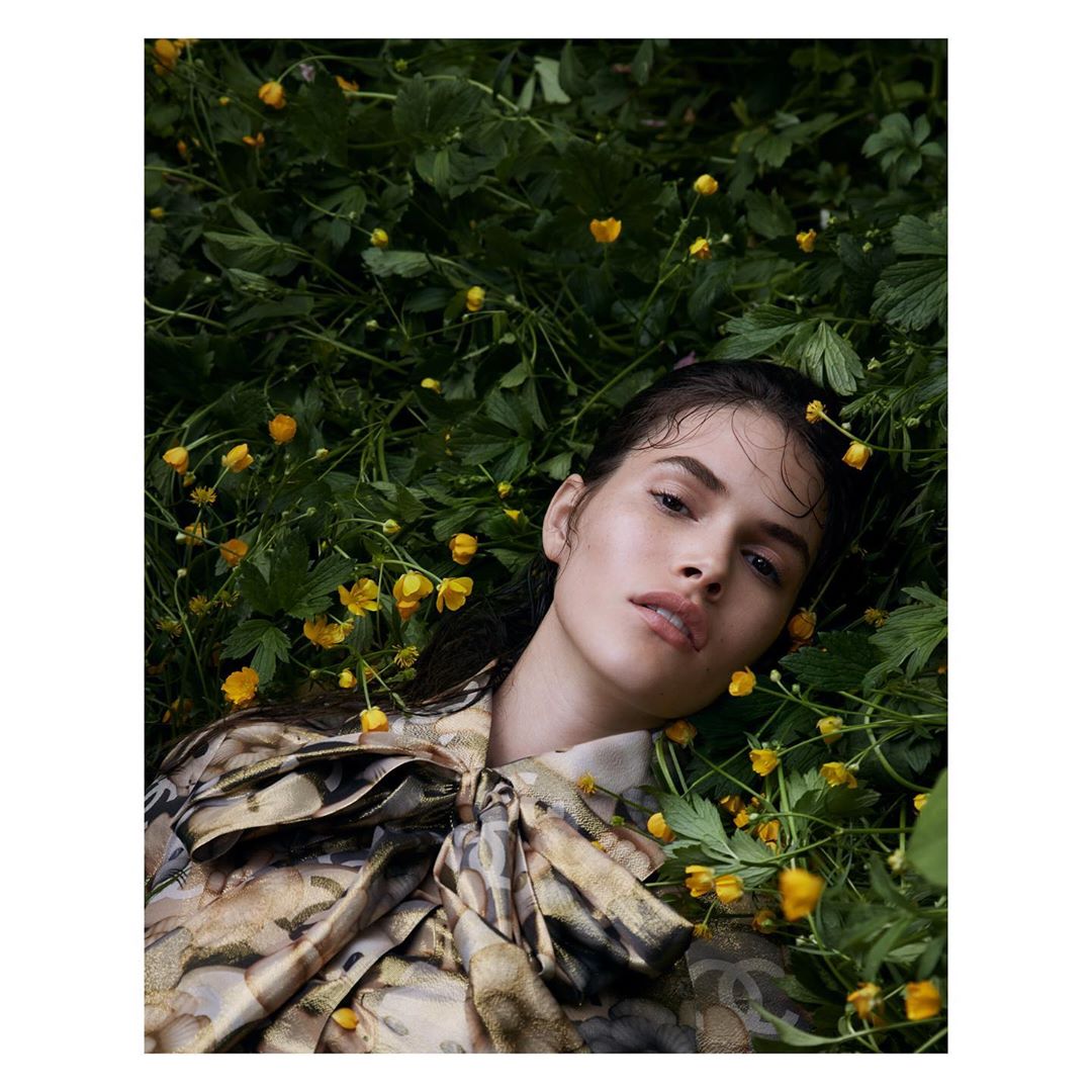 Brunette woman in a patterned blouse lying among green foliage and yellow flowers. Close-up shot, shallow depth of field, outdoor environment.