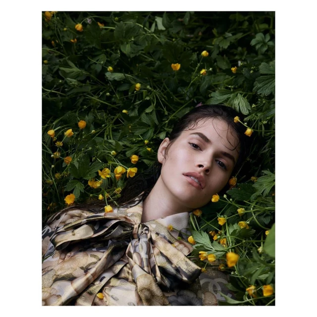 Brunette woman in a patterned blouse lying among green foliage and yellow flowers. Close-up shot, shallow depth of field, outdoor environment.