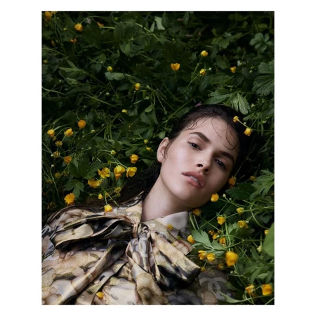 Brunette woman in a patterned blouse lying among green foliage and yellow flowers. Close-up shot, shallow depth of field, outdoor environment.