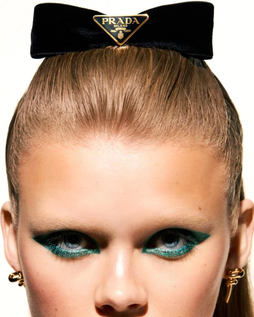 Close-up of a woman's face with blue eyes and glittery green eyeliner wearing a black velvet Prada bow headband with logo.