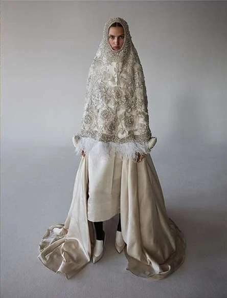 Full-length shot of a fair-skinned young woman wearing an ornate beaded cape with white trim, a satin skirt, and white pointed shoes.