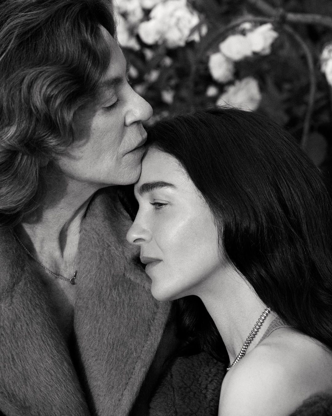 Intimate black and white portrait of two women in soft focus with a rose bush backdrop, showcasing a tender moment in natural light.