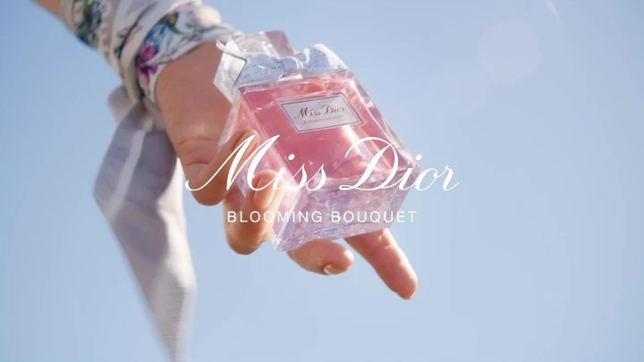 Hand holds a bottle of Miss Dior Blooming Bouquet perfume against a clear blue sky, showcasing a fashion and beauty product.