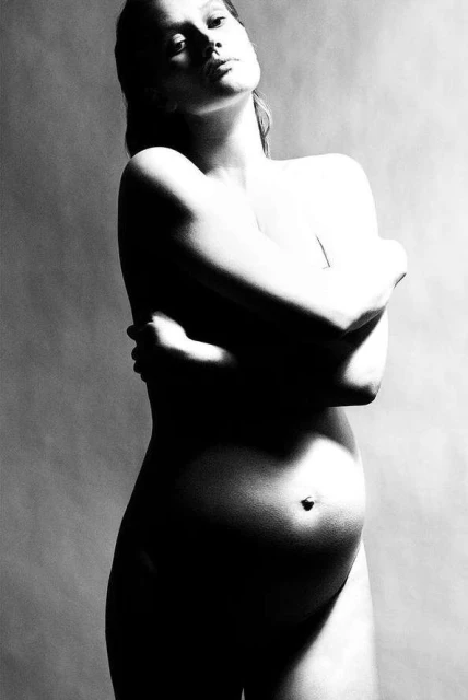 Monochrome portrait of a pregnant woman, nude and embracing herself, against a neutral backdrop, captured in a studio with soft lighting.