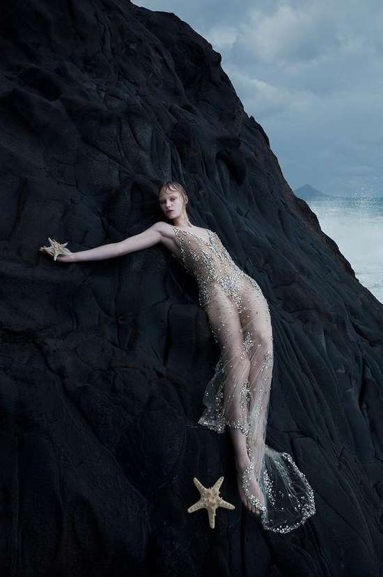 Woman in beaded gown poses with starfish on dark rocks by the ocean, portrait orientation. Fashion, beauty, and nature elements.