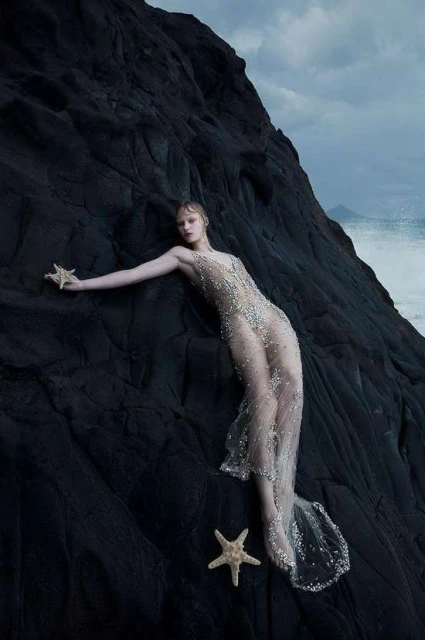 Woman in beaded gown poses with starfish on dark rocks by the ocean, portrait orientation. Fashion, beauty, and nature elements.