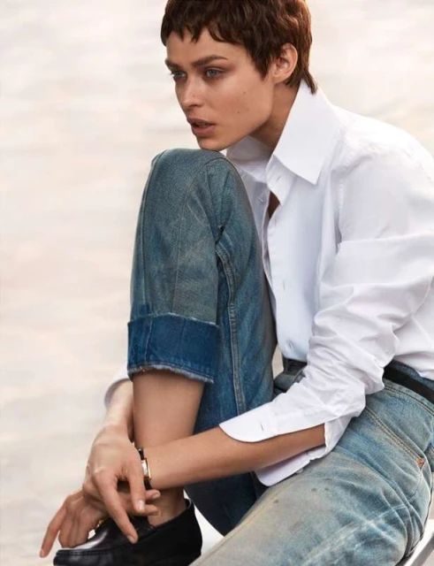 A young adult woman with short hair poses while wearing a white collared shirt, blue jeans, and black shoes, against an out-of-focus backdrop.