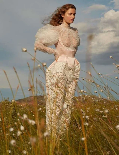 A woman poses in a white lace dress with puffy sleeves in an outdoor field under a blue sky with light cloud cover and low mountain.