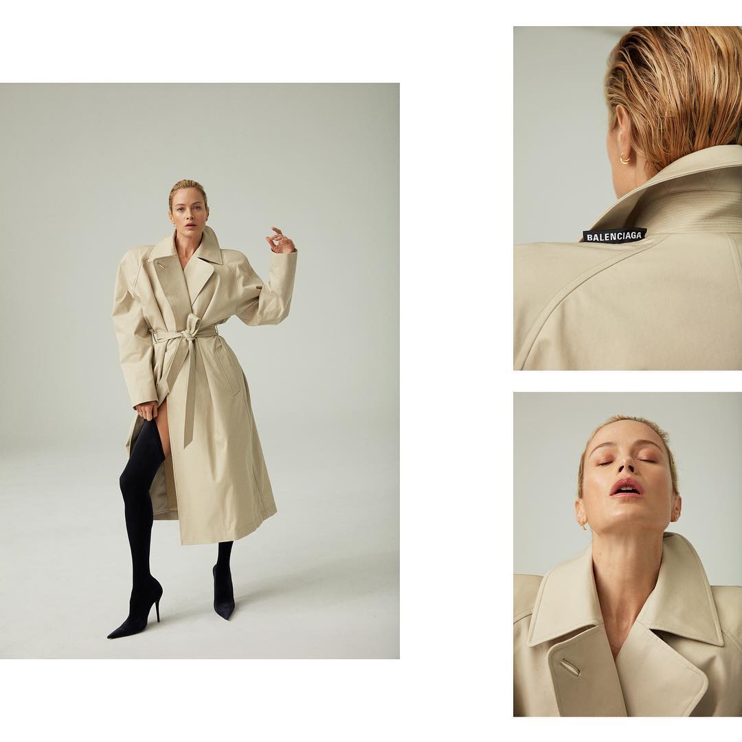 Full-length shot of a blonde woman wearing a beige trench coat, black hosiery, and pointed-toe heels against a neutral backdrop.
