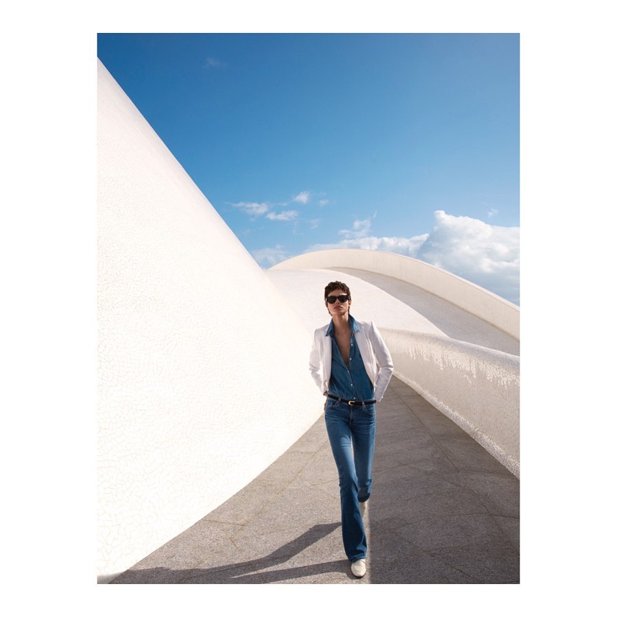 A woman walks in front of a modern white building under a blue sky, wearing jeans, a denim shirt, and a white jacket.