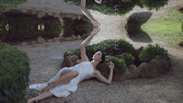 Woman in a white dress lies on gravel with mirrored reflection, creating a surreal and ethereal effect in an outdoor garden setting.
