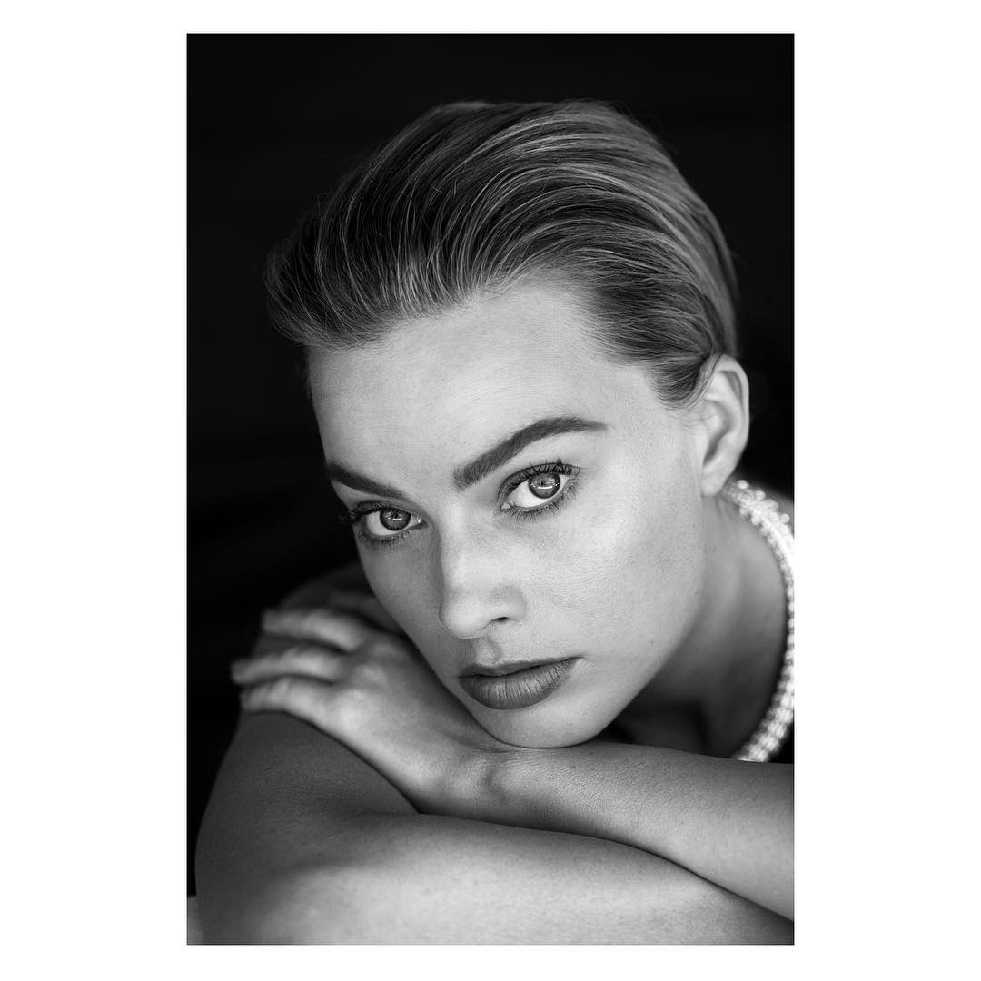 Striking black and white portrait of a young woman with her arms crossed, featuring an elegant pearl necklace against a dark backdrop.