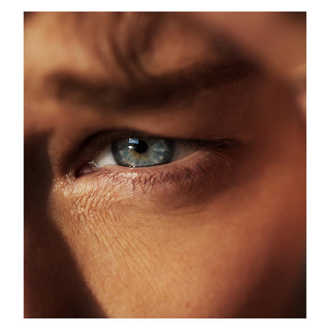 Macro close-up portrait of a young adult male's light blue eye and surrounding skin in warm lighting, centered in a square format.