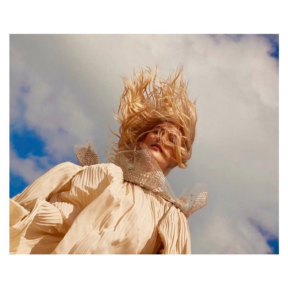 Blonde woman with hair styled upward, wearing a flowing, pleated dress with a beaded collar, set against a blue sky with white clouds.