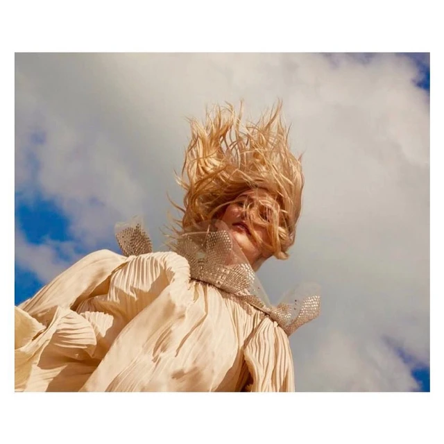 Blonde woman with hair styled upward, wearing a flowing, pleated dress with a beaded collar, set against a blue sky with white clouds.