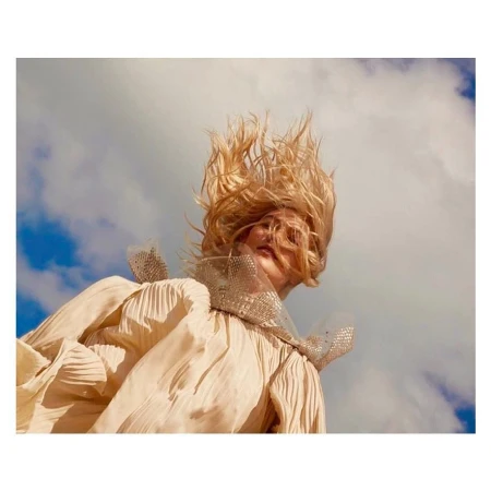 Blonde woman with hair styled upward, wearing a flowing, pleated dress with a beaded collar, set against a blue sky with white clouds.