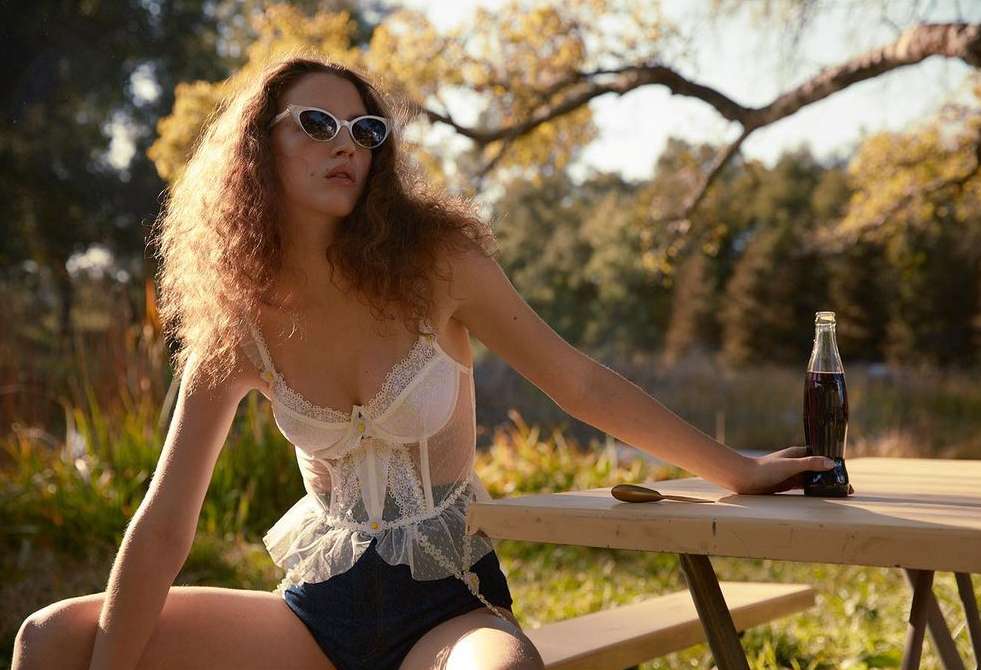A woman in lingerie poses on a picnic table outdoors with a bottle of soda, wearing sunglasses and backlit by sunlight.