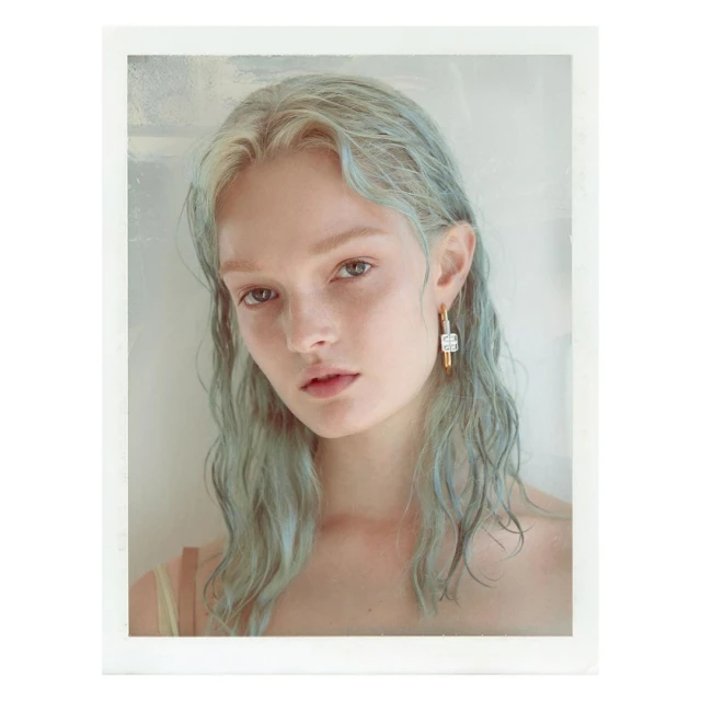 Close-up portrait of a young woman with wavy, blue-tinted hair, wearing gold and silver earrings against a light background in a studio.