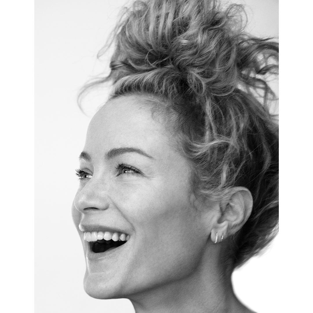 Portrait of a cheerful woman with an updo hairstyle and earrings, smiling broadly against a bright, neutral background in black and white.