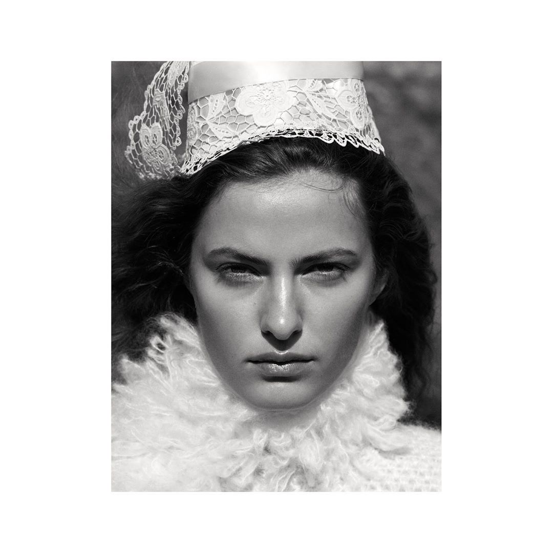 Monochrome portrait of a woman with a serious expression wearing a white lace headpiece and ruffled collar against a blurry background.