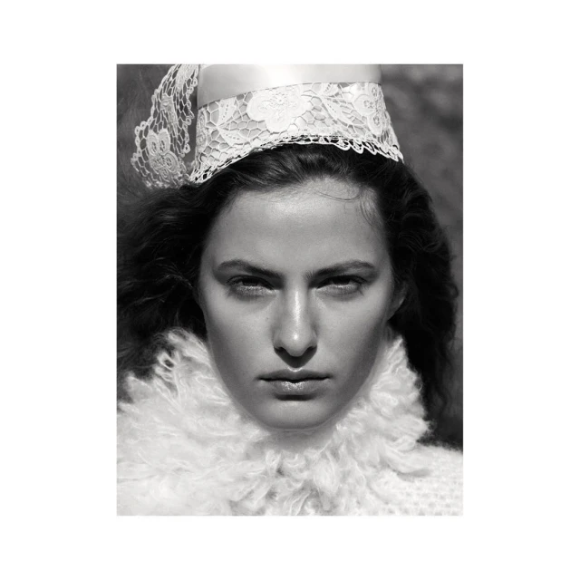Monochrome portrait of a woman with a serious expression wearing a white lace headpiece and ruffled collar against a blurry background.