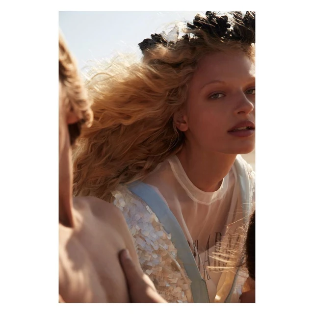 Fashion shot of a blonde woman with a feathered headpiece looking at the camera, with fair-skinned people blurred in the background.