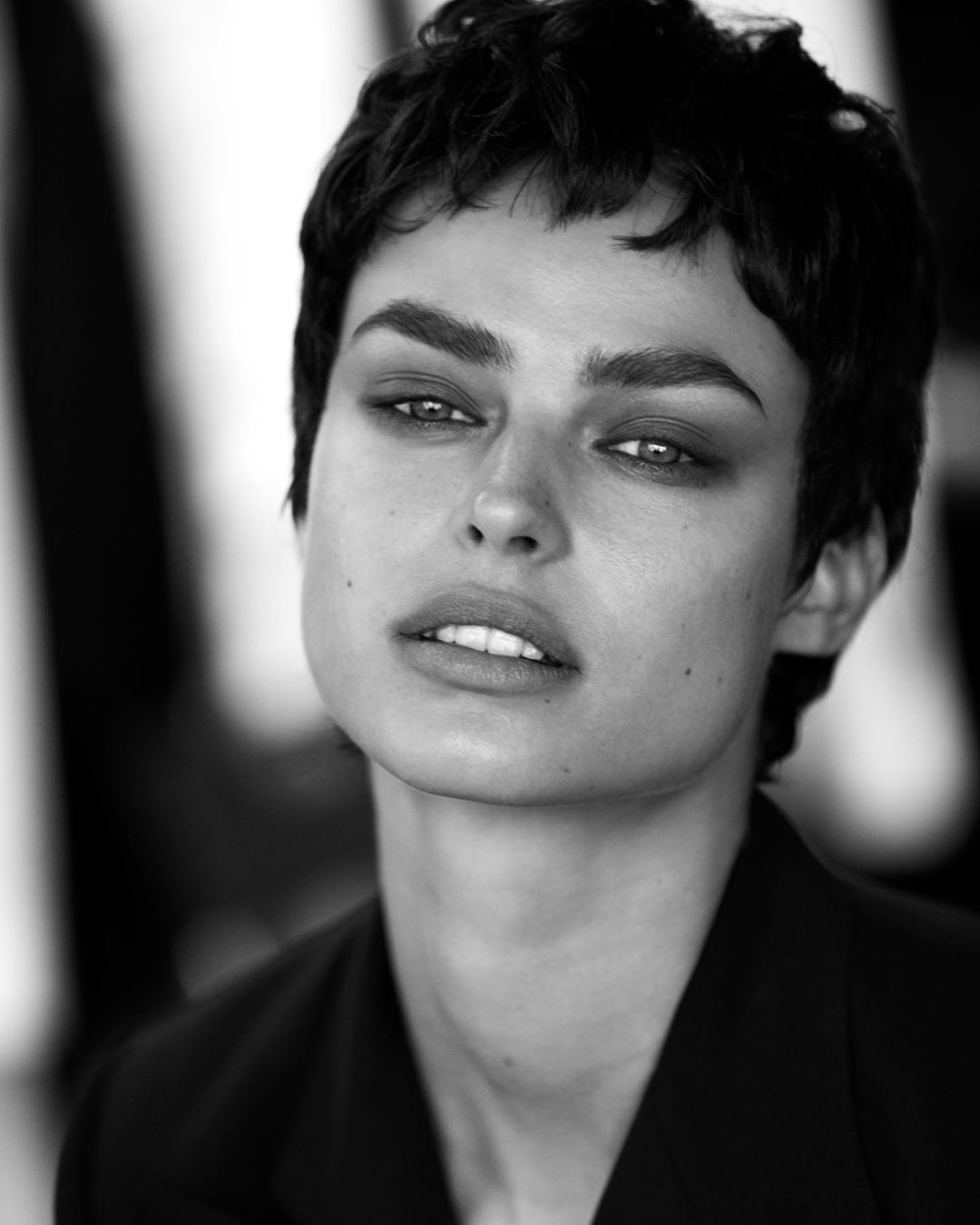 Striking black and white portrait of a woman with short hair, a defined jawline, and intense gaze, wearing a dark blazer against a neutral background.