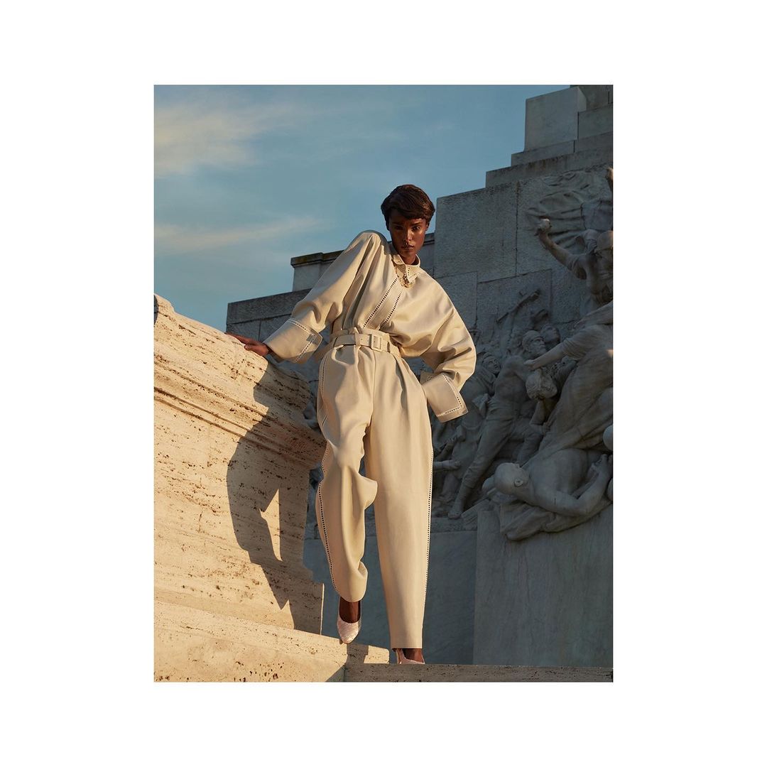 Model wearing a belted cream jumpsuit standing on stone steps outdoors against a monumental carved relief wall.