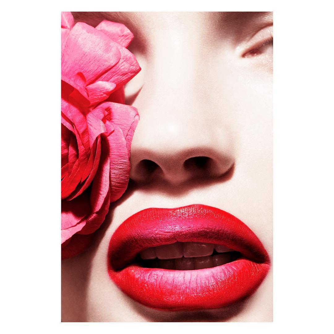 Close-up portrait of a fair-skinned woman with vibrant red lipstick, a pink flower framing her face, and soft, diffused lighting.
