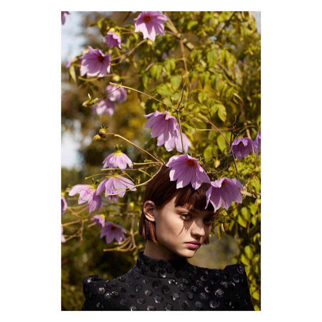 Portrait of a young woman with short brown hair and a patterned top, framed by large, light purple flowers and lush green foliage.