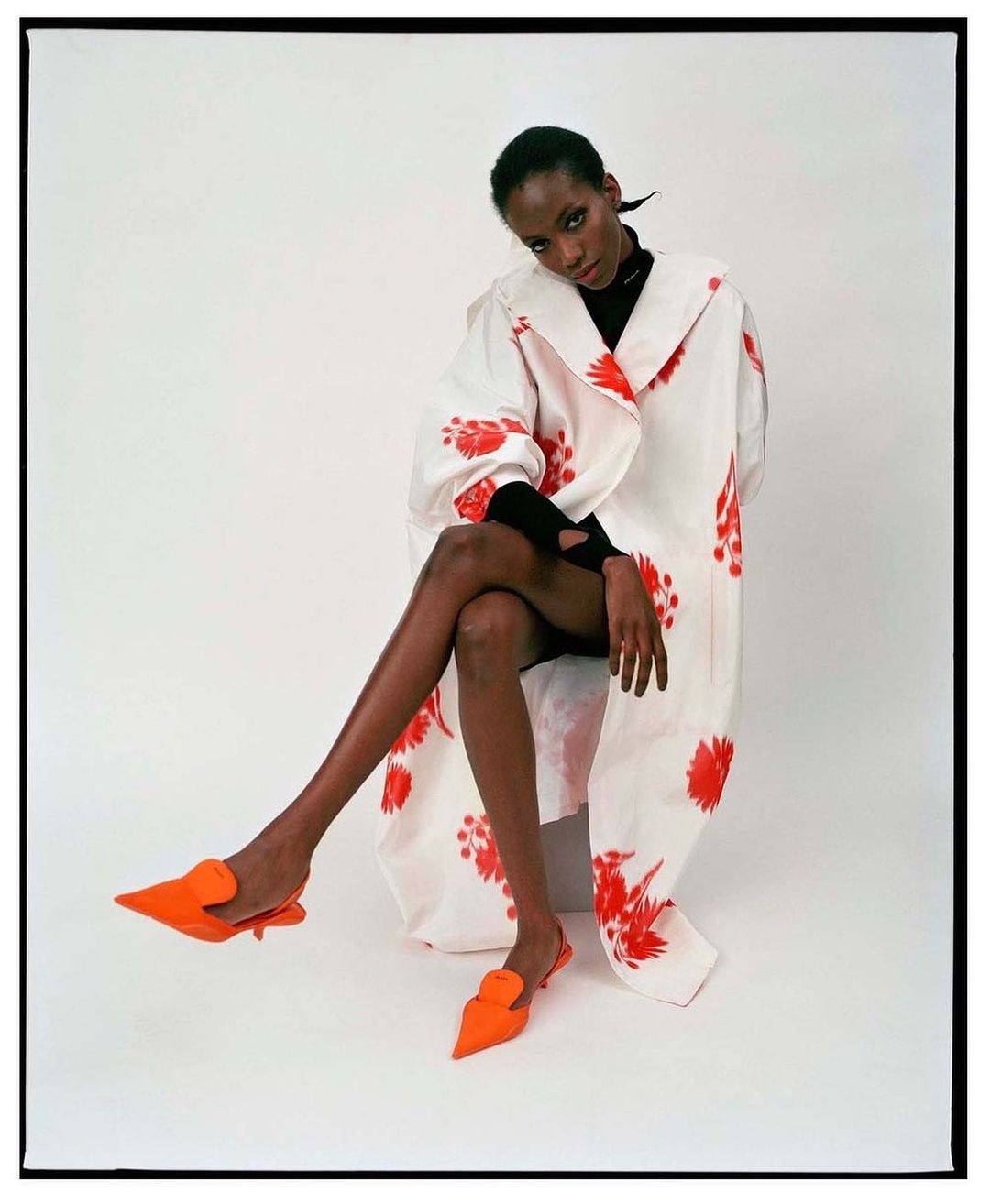 A single black woman sits with legs crossed, wearing a white floral-print coat and orange shoes in a brightly lit photo studio.
