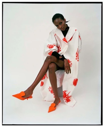 A single black woman sits with legs crossed, wearing a white floral-print coat and orange shoes in a brightly lit photo studio.