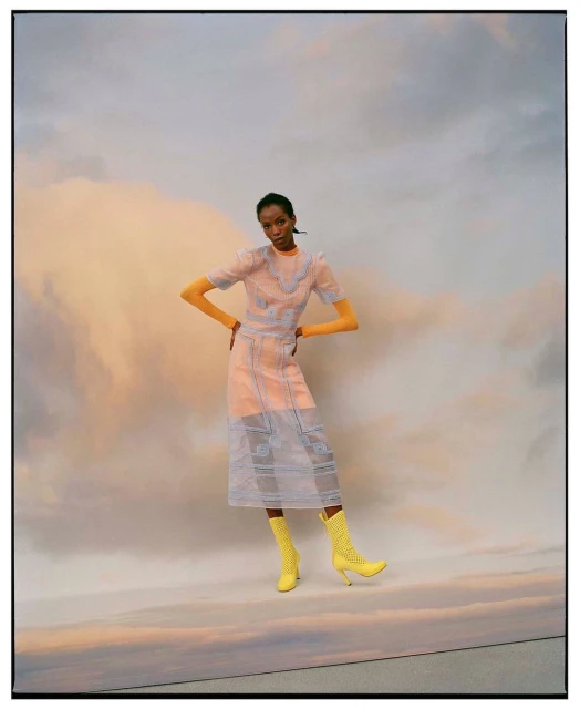Elegant Black woman in a sheer patterned dress and yellow boots poses against a cloudy backdrop in a fashion editorial-style shot.