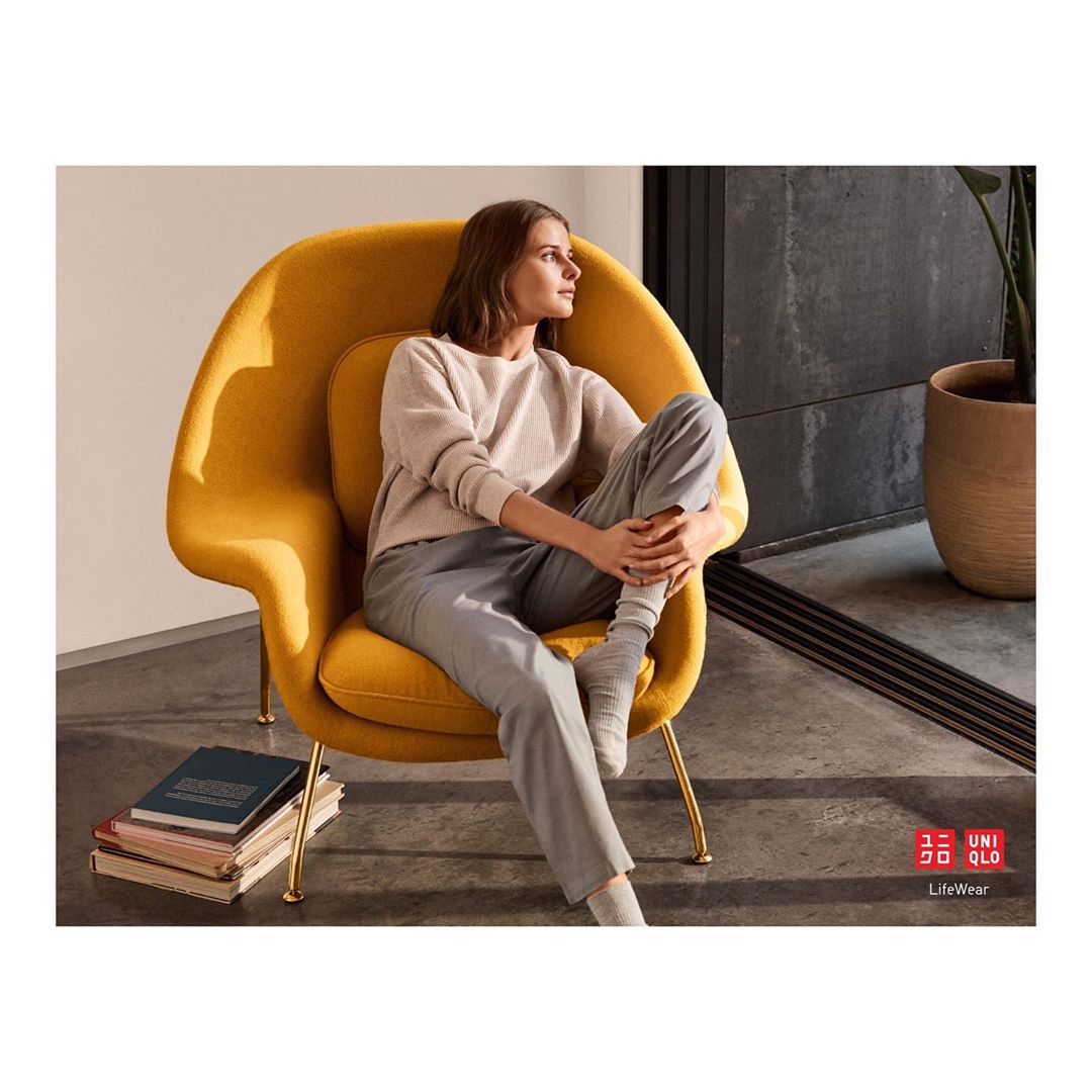 Indoor shot of an adult woman in loungewear sitting in a yellow chair, looking out of frame, with books stacked near the chair.
