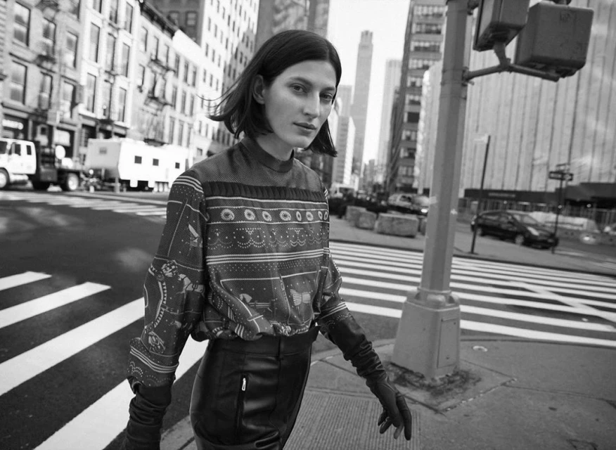 Black and white portrait of an adult female in an urban setting. She is wearing a patterned top, long gloves and a skirt, on a city street.