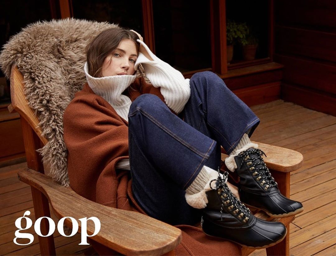 A woman in a white sweater and jeans sits in a wooden chair with a fur blanket, wearing lace-up black boots in an indoor setting.