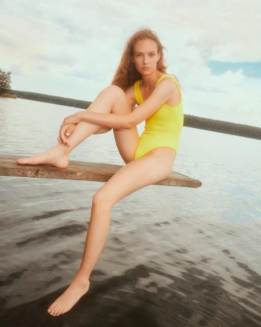 A young, slender woman in a yellow swimsuit sits on a wooden plank above a calm lake, with a forest visible in the background.