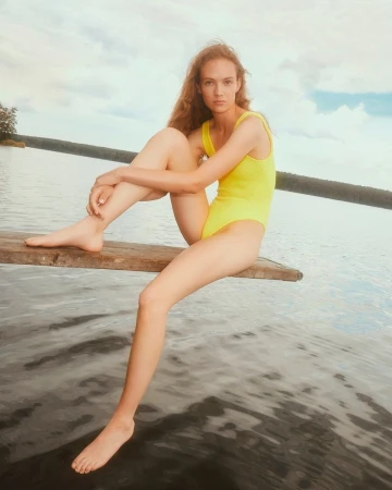 A young, slender woman in a yellow swimsuit sits on a wooden plank above a calm lake, with a forest visible in the background.