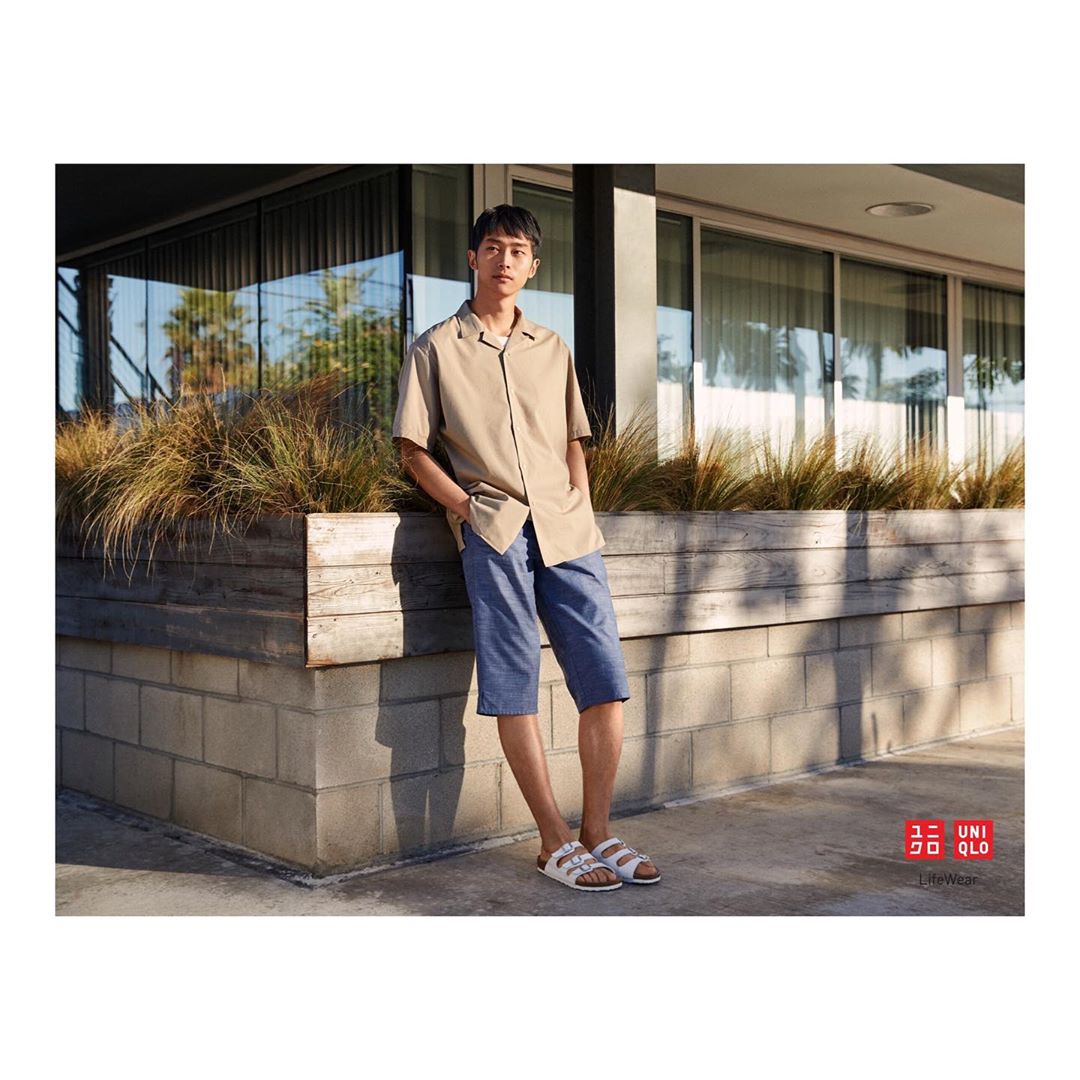 A young adult man stands outdoors in a beige shirt and blue shorts, casually posed against a textured wall, showcasing a contemporary style.