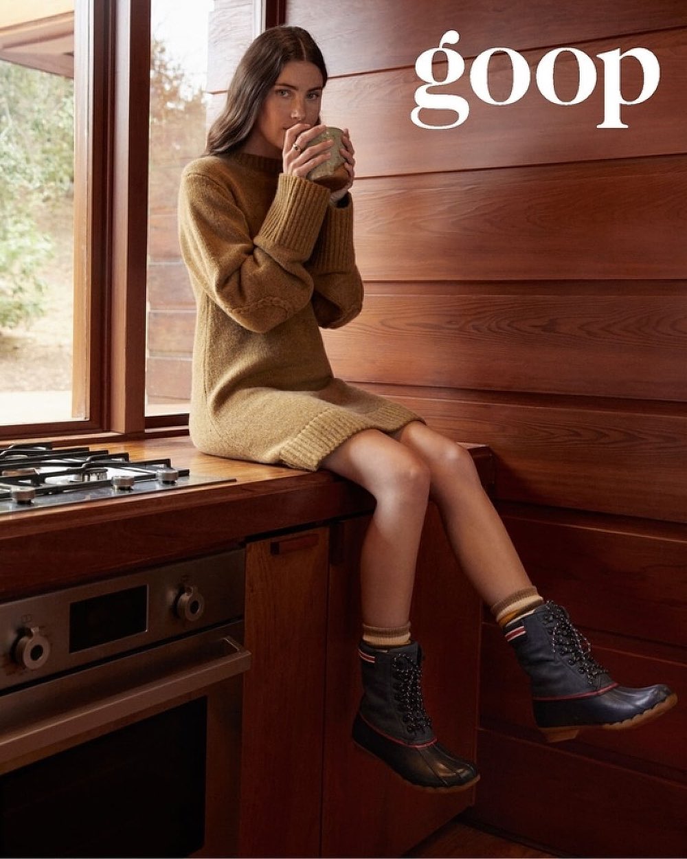A young woman in a knit dress and duck boots sits on a kitchen counter drinking from a mug, with wooden cabinets in the background.