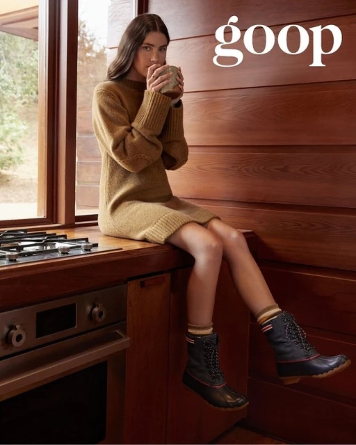 A young woman in a knit dress and duck boots sits on a kitchen counter drinking from a mug, with wooden cabinets in the background.