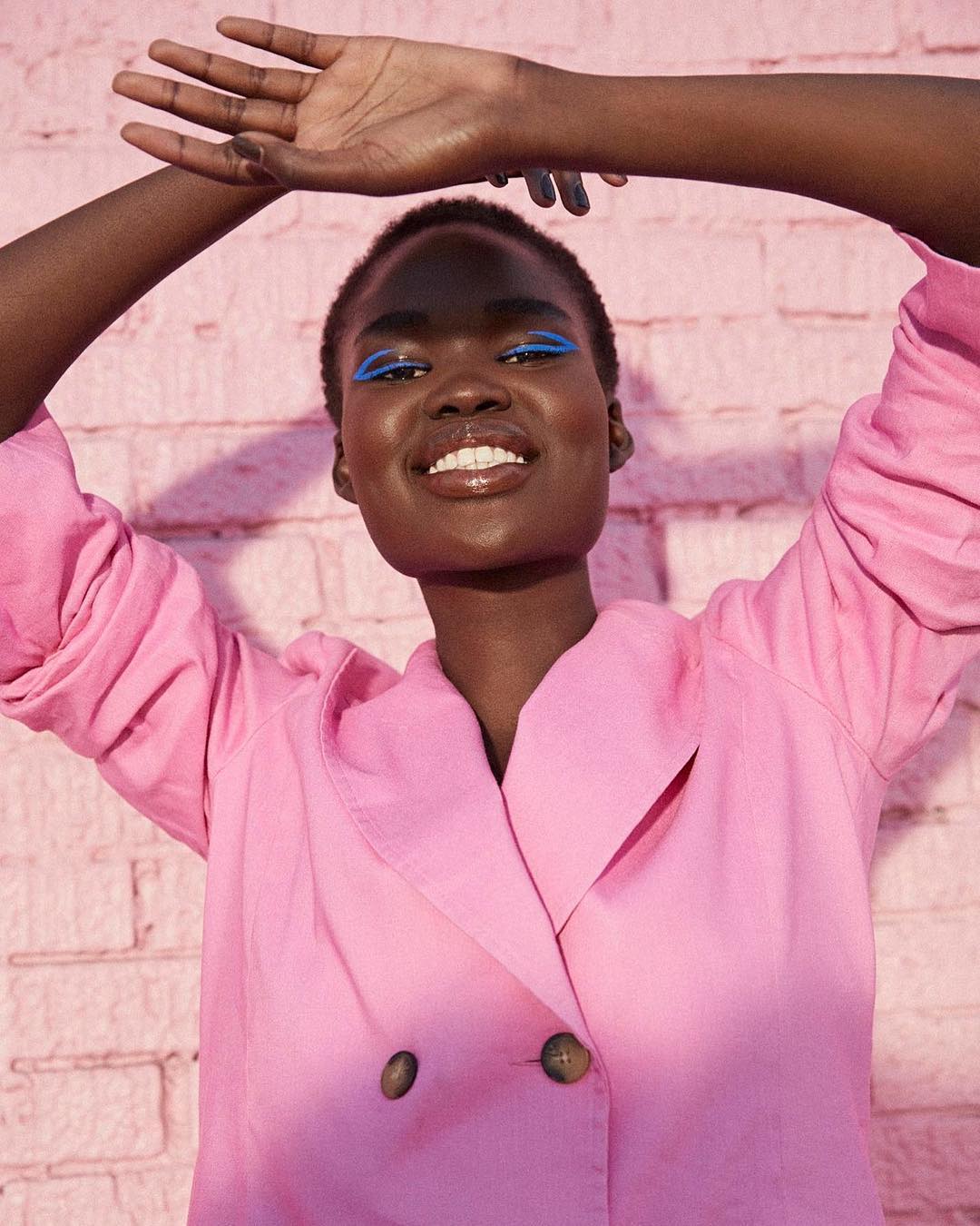 Smiling African American woman with blue eyeliner posing in front of a pink brick wall wearing a pink jacket, shot in natural lighting.